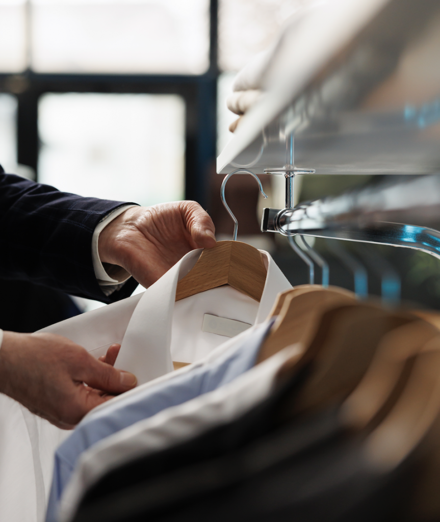 Nahaufnahme: Eine Kleiderstange mit mehreren Anzugshemden. Eine Hand hängt gerade ein Hemd daran auf.“